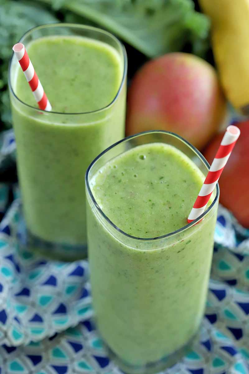 Vertical oblique shot of two tall glasses filled with green smoothie and striped red and white straws, on a dark and light blue patterned cloth, with fresh produce in the background.