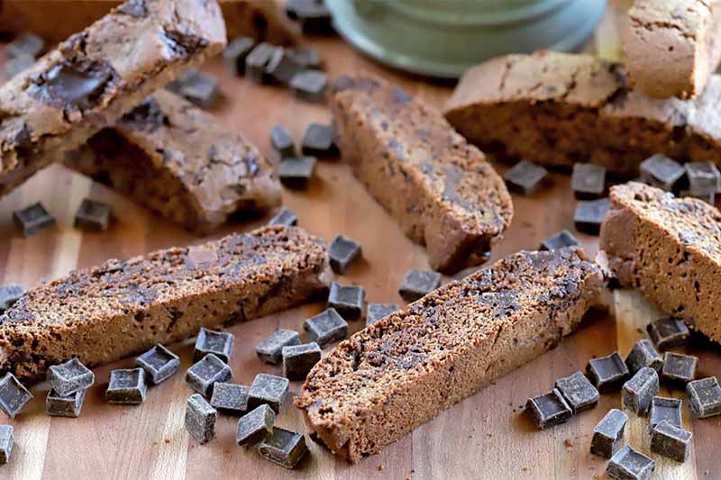 Homemade biscotti and scattered chocolate chunks on a wood surface, with a jade green ceramic mug to the right in the background.