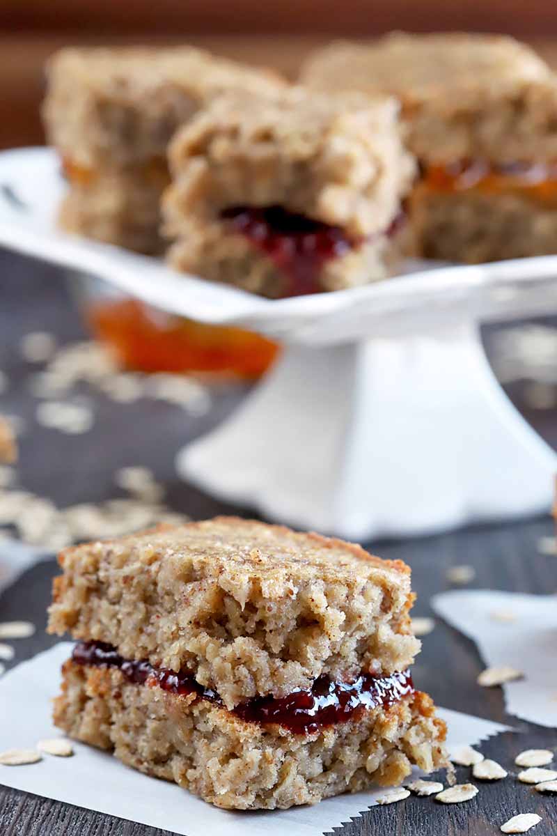 Closeup of an oat sandwich-style breakfast bar with red jelly in the middle, on a square piece of white parchment paper, with a white pedestal-style serving dish in the background with more of the bars on top, and scattered uncooked oats around the base, on a dark brown wood surface.