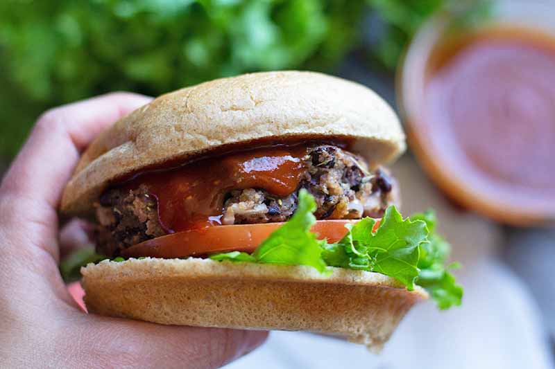 Horizontal close-up image of a hand holding a whole patty with bread, sauce, lettuce, and tomatoes.