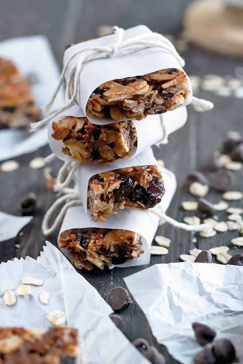 Vertical image of a stack of four homemade granola bars wrapped in paper and tied with string, on a dark brown wood surface with more bars on rectangular pieces of parchment paper, with scattered oats and chocolate chips.
