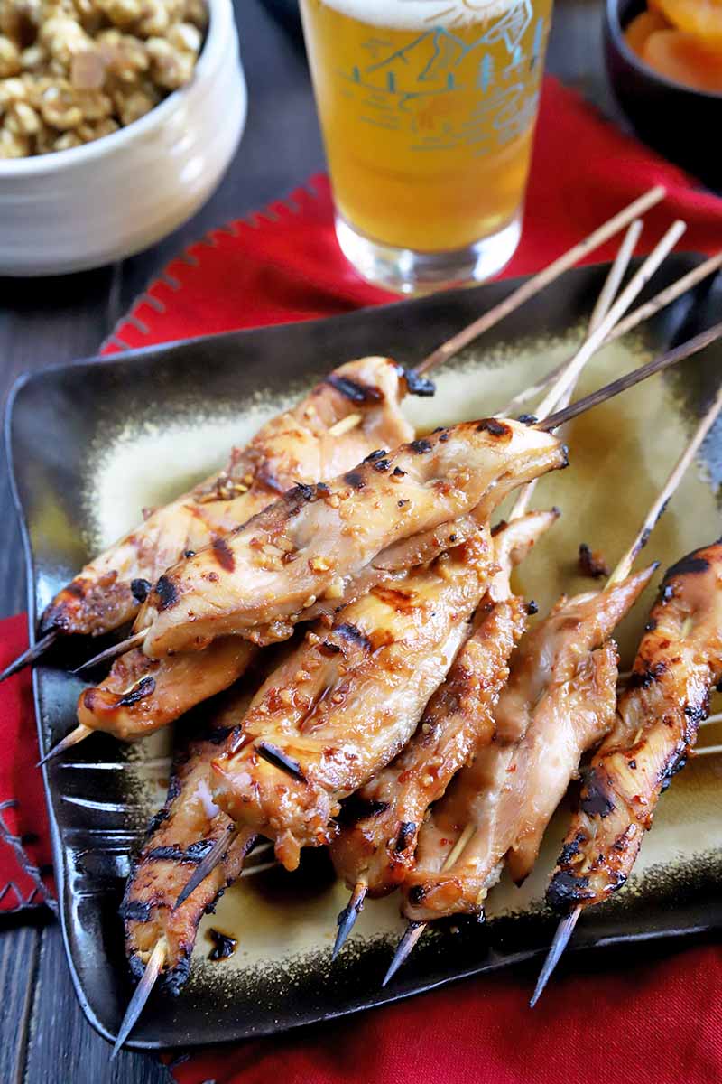 Vertical image of a square black and gold plate of grilled marinated Japanese-style chicken on bamboo skewers, on a red cloth with a glass of beer and bowls of snack foods in the background, on a wooden table.