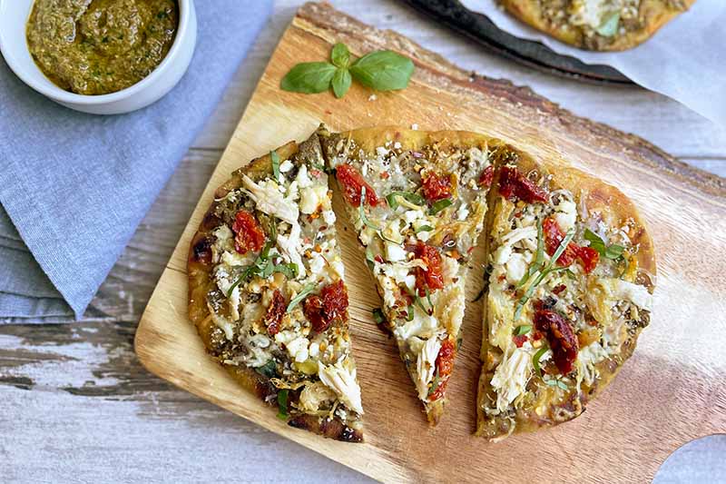 Horizontal image of three slices of an oblong pizza on a wooden cutting board next to pesto and a blue towel.