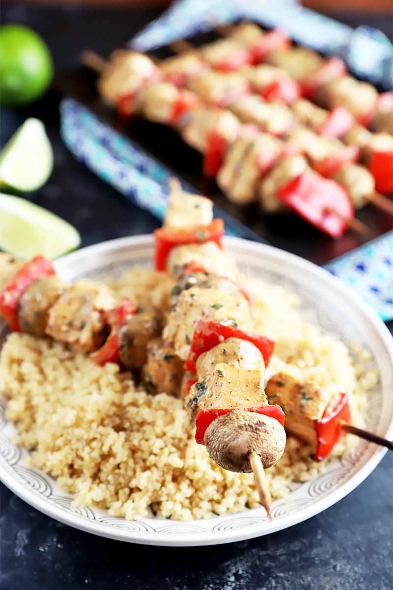 Vertical image of two halibut kebabs over a plate of cooked grains, with a black platter and lime wedges in the background.