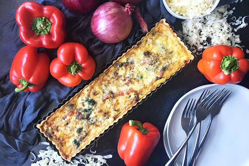 Horizontal image of a whole tart topped with melted cheese on a dark surface next to forks, plates, cheese, and assorted vegetables.