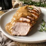 Horizontal image of shingled slices of seared meat topped with gravy on a white plate next to herbs and tan towels.