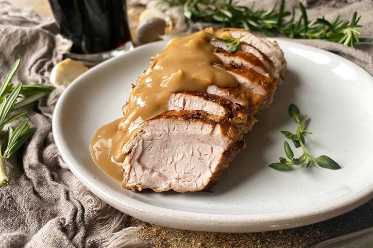 Horizontal image of shingled slices of seared meat topped with gravy on a white plate next to herbs and tan towels.