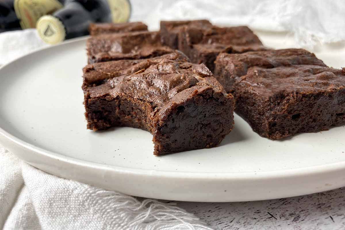Horizontal image of individual brownies, one with a large bite, on a white plate with bottles in the background.