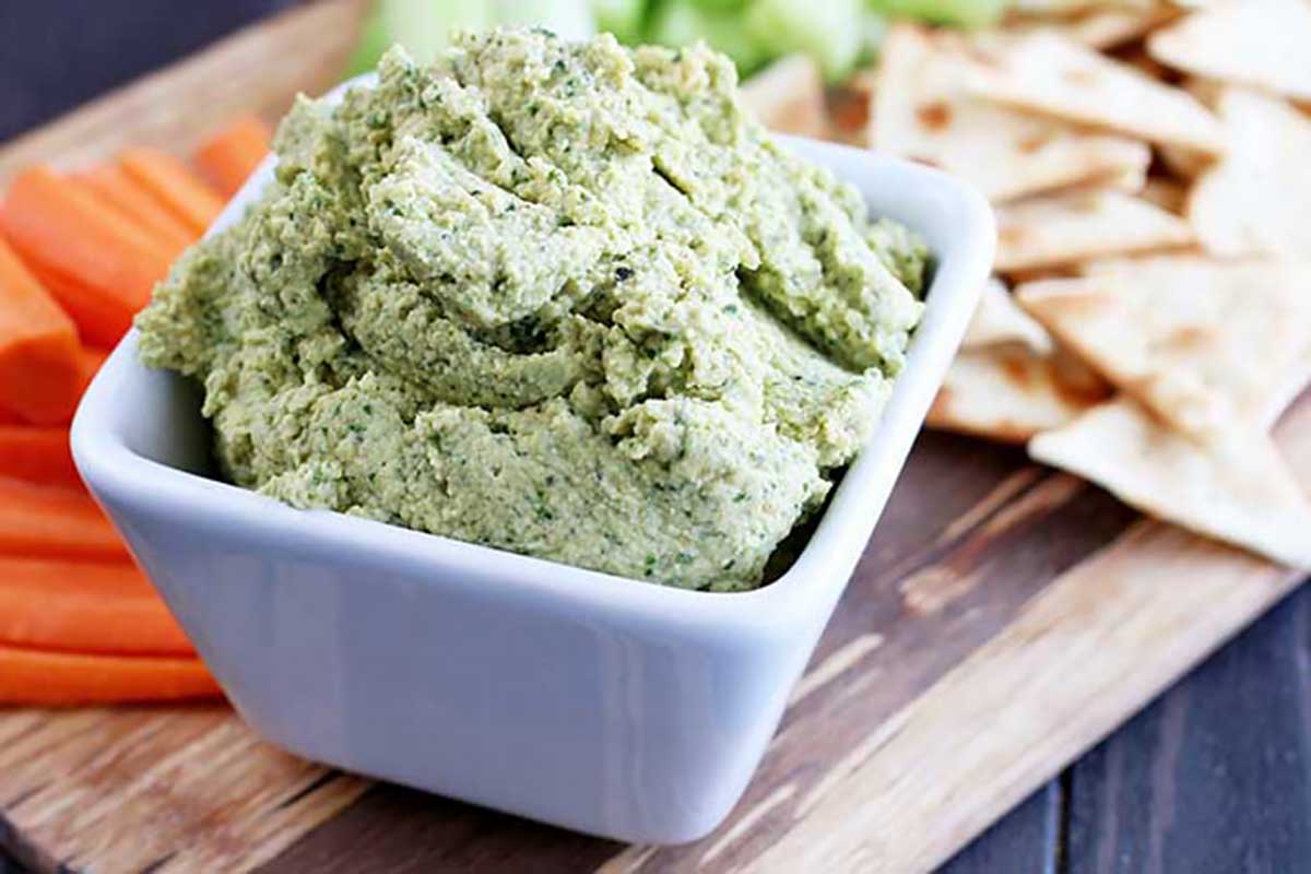 Horizontal image of a kale and basil bean puree in a small ramekin on a wooden platter next to pita, carrot, and celery.