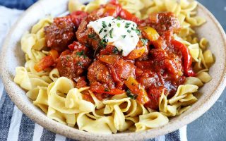 Horizontal image of a tan bowl filled with egg noodles underneath a hearty mound of sauce garnished with a dollop of sour cream and chopped herbs.