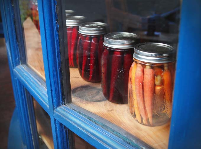 A row of carrots and parsnips pickled in mason jars is displayed in a glass window cabinet with blue frames.
