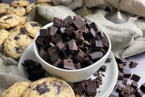 Horizontal image of a bowl filled with dark brown chunks of candy on a plate next to a tan towel and cookies.