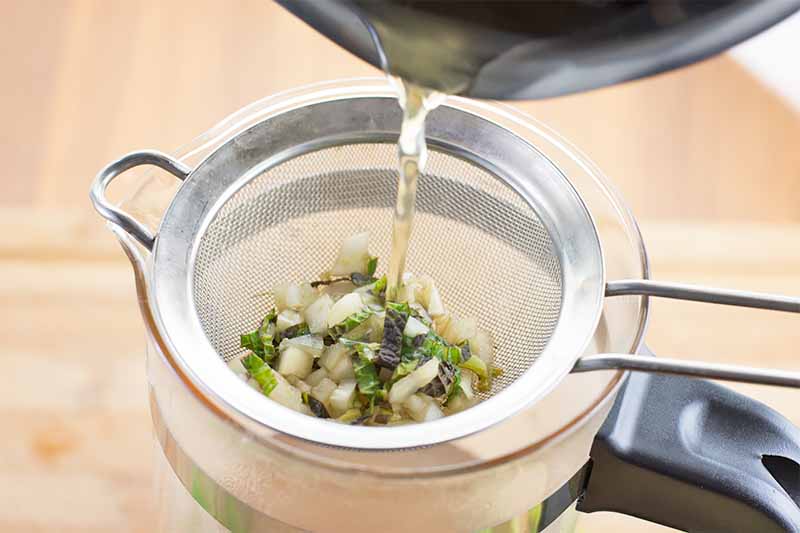Horizontal image of straining a liquid with chopped ingredients in a small metal strainer over a liquid measuring cup.