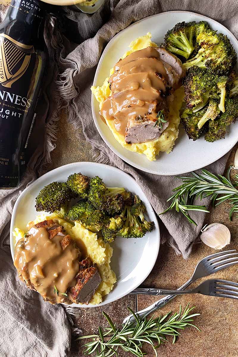 Vertical top-down image of shingled slices of meat on top of mashed potatoes and broccoli, set up on tan towels next to a bottle, herbs, and forks.