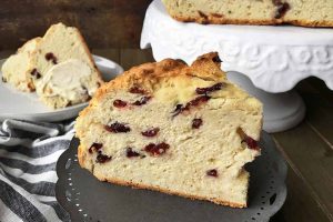 Horizontal image of an upright slice of Irish soda bread on a gray stand, with a white stand and more baked goods in the background.