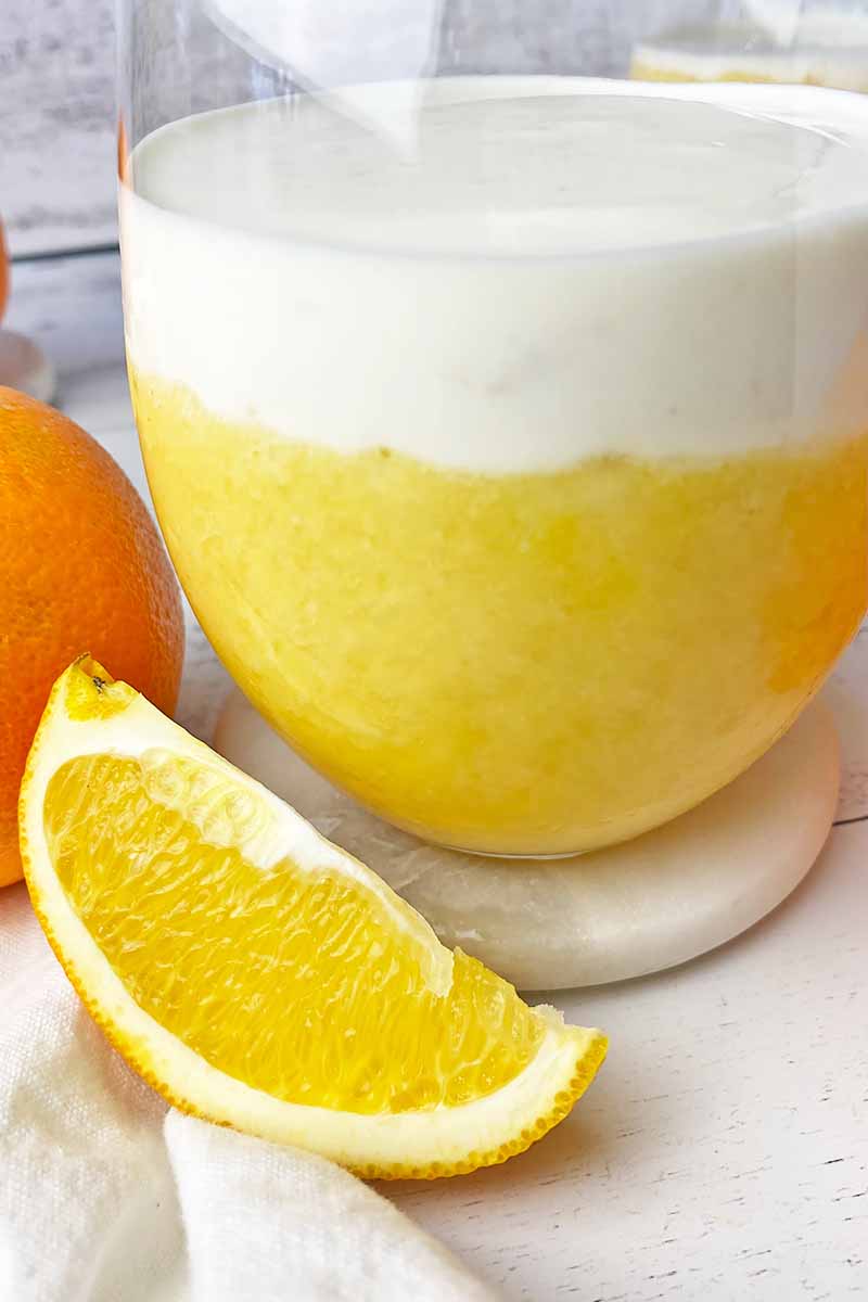 Vertical close-up image of layers of a beverage in a glass next to a slice of fruit.