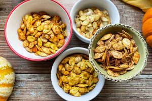 Horizontal image of four bowls filled with assorted spiced pumpkin seeds on a wooden surface.