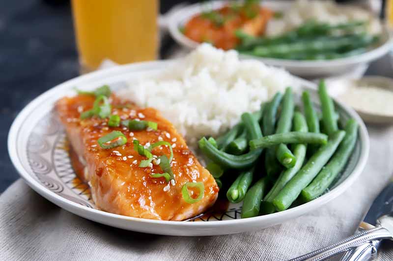 Horizontal image of two white plates with green beans, rice, and fish fillets topped with green onions and sesame seeds.