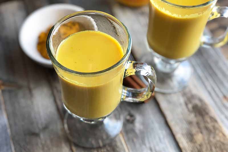 Horizontal image of two glass mugs with handles filled with a bright orange mixture in front of a bowl with ground cinnamon.