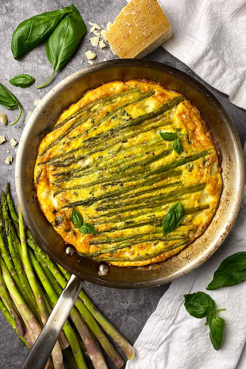 Vertical top-down image of a whole frittata in a skillet with asparagus spears and basil leaves.