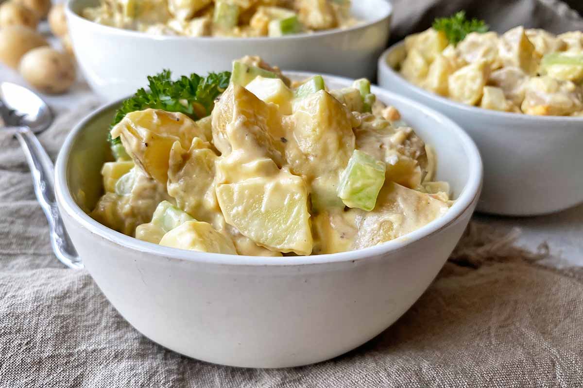 Horizontal close-up image of small bowls filled with a potato salad on a tan towel.