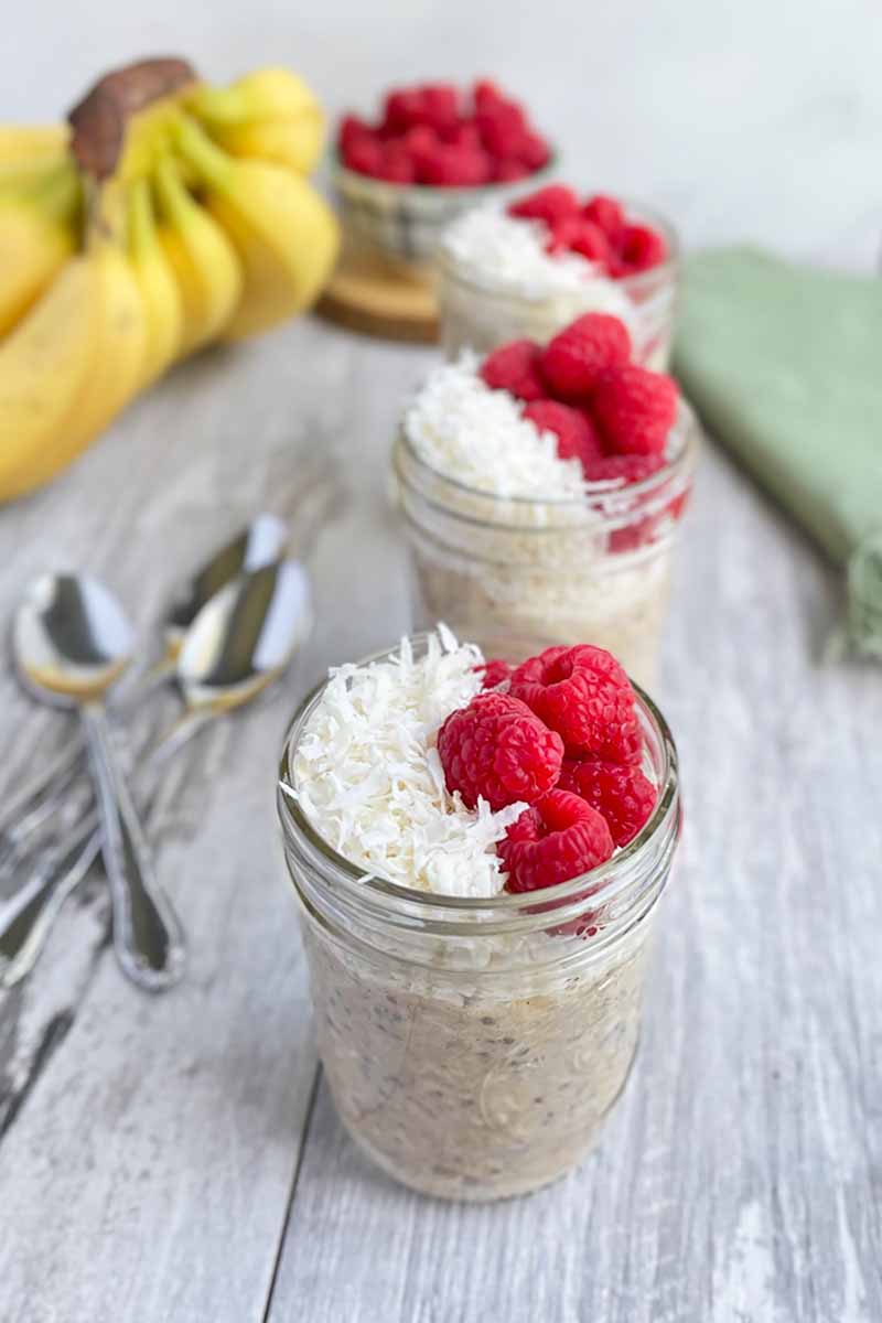 Vertical image of multiple mason jars filled with oatmeal topped with coconut shreds and raspberries, with bananas in the background.