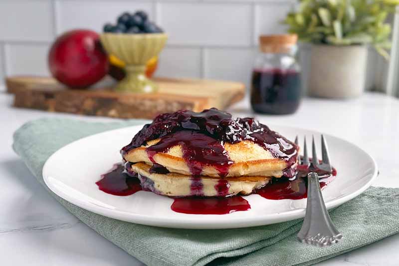 Horizontal image of pancakes on a white plate with a purple sauce poured over them.