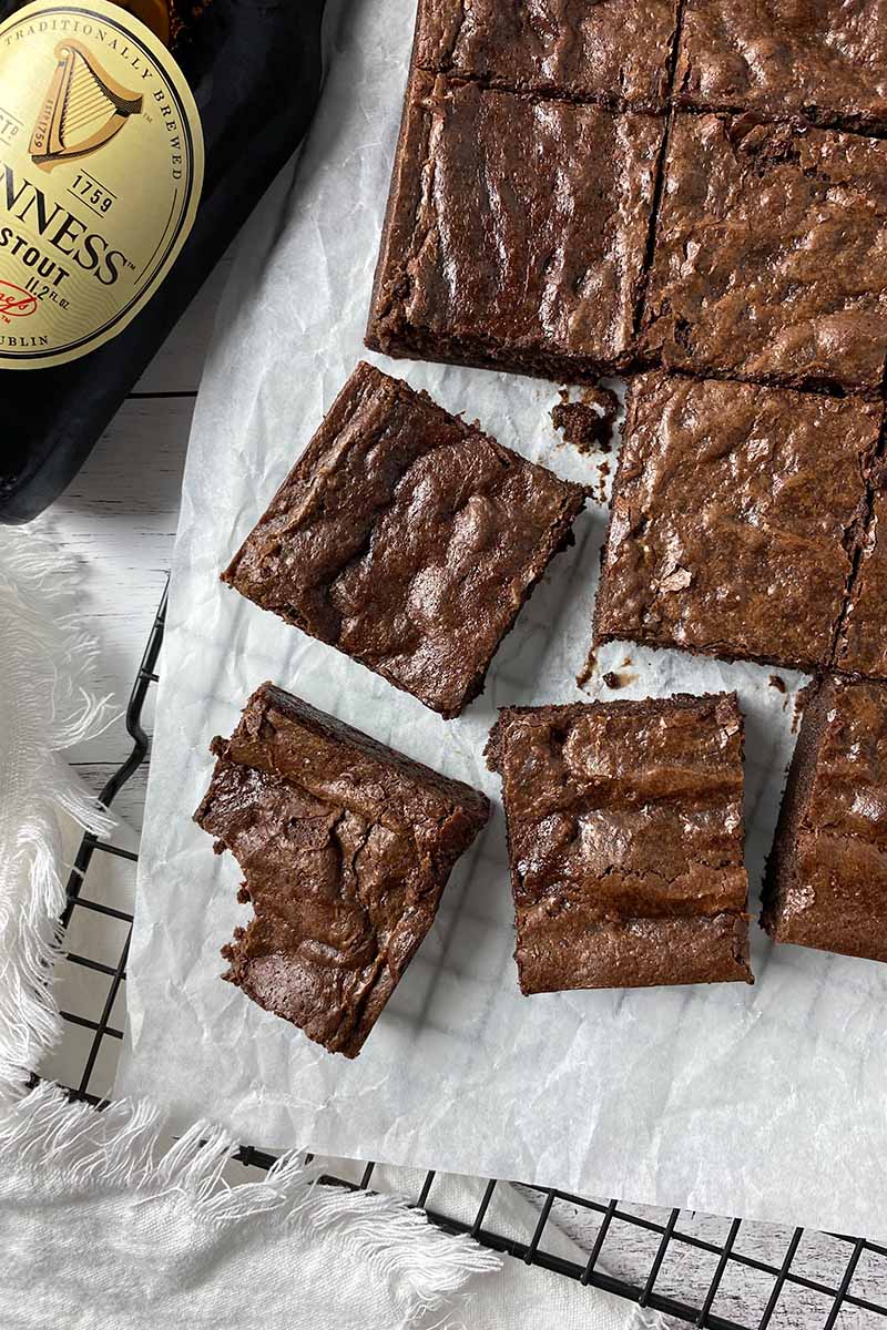 Vertical top-down image of portioned individual square baked goods on a parchment paper next to bottles Guinness.