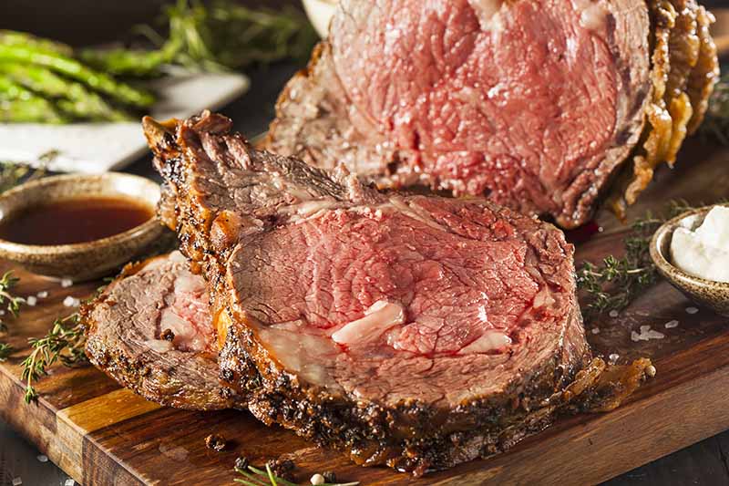 Horizontal image of a thick slice of rare meat on a wooden cutting board next to sauces in bowls.