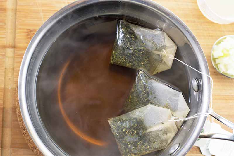 Horizontal image of a pot filled with hot water and tea bags steeping in it on a wooden surface.