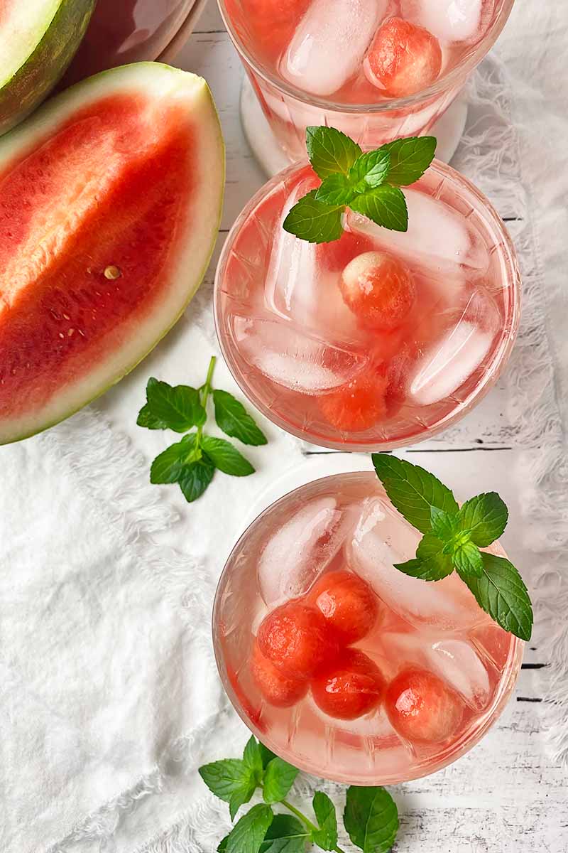 Vertical top-down image of a neat row of three tumblers filled with a beverage with fruit balls, ice cubes, and fresh herb leaves for a garnish next to white towels.