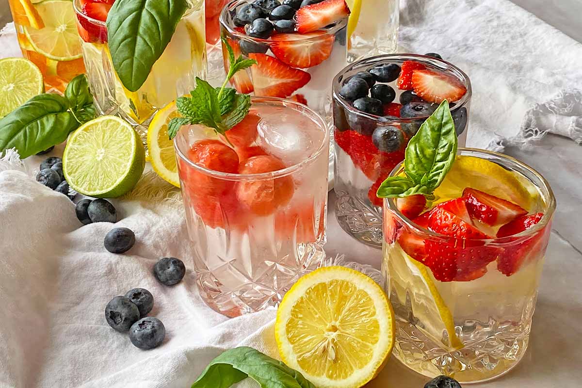 Horizontal image of assorted beverages in glasses next to fruit, herbs, and white towels.