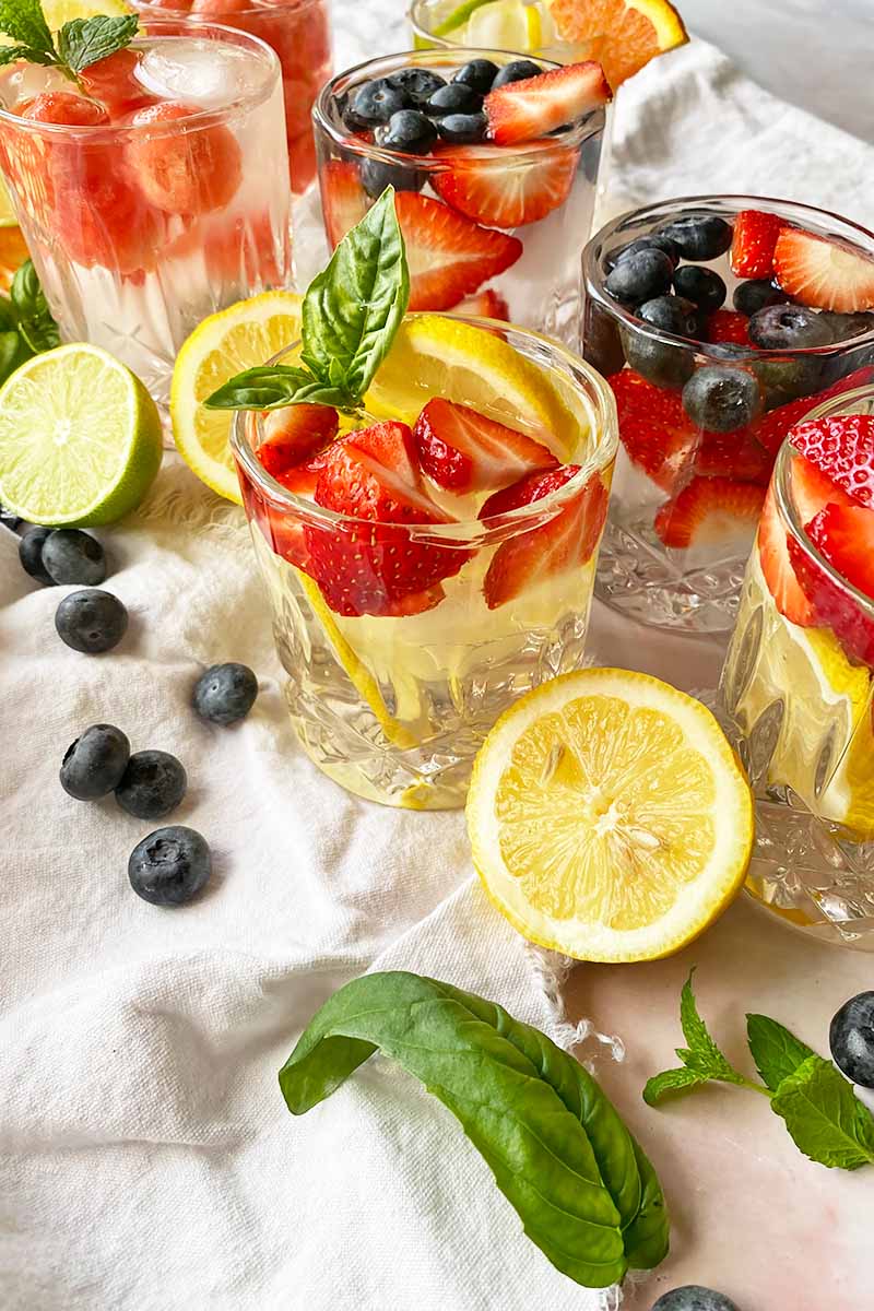 Vertical image of assorted beverages with garnishes in glasses on white towels next to fresh fruits and herbs.