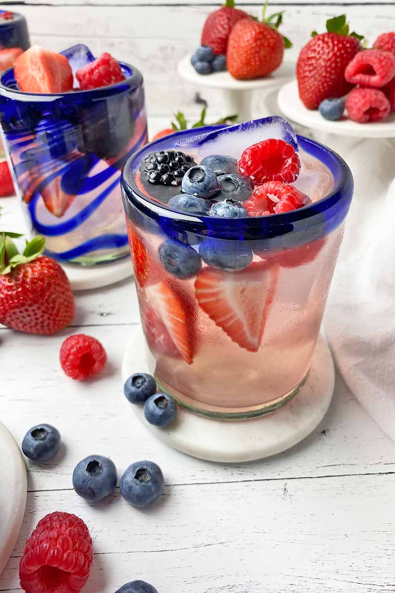 Vertical image of blue-rimmed glasses filled with a light pink beverage and assorted fresh garnishes on white coasters in front of small white cake stands.