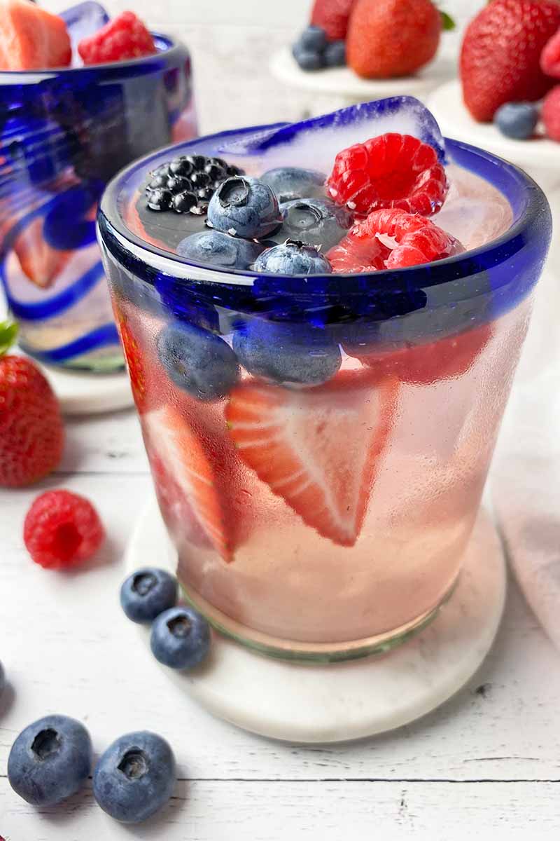 Vertical close-up image of blue-rimmed glasses filled with a light pink beverage and assorted fresh garnishes on white coasters.