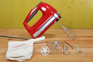 KitchenAid KHM926 9-Speed Hand Mixer with the blending, mixing beaters, wire whisk, and bread hook attachments on a maple butcher block table and lime green wall background.