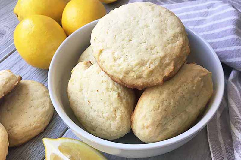 Horizontal image of a white bowl with a pile of freshly baked fluffy cookies surrounded by three whole lemons, lemon wedges, and a striped gray towel on a gray wooden surface.