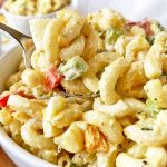 Horizontal close-up image of a spoonful of a chilled and creamy elbow macaroni dish in a white bowl with assorted vegetables.