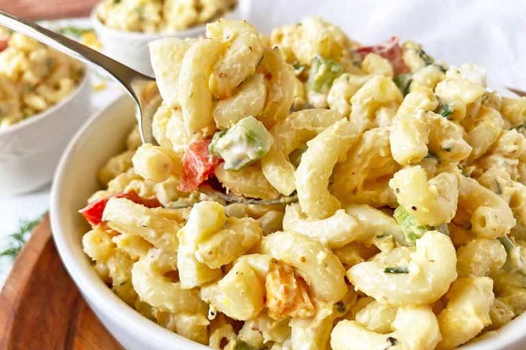 Horizontal close-up image of a spoonful of a chilled and creamy elbow macaroni dish in a white bowl with assorted vegetables.