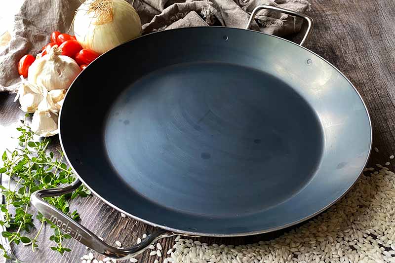 Horizontal image of an empty shallow cookware with handles next to produce and herbs.