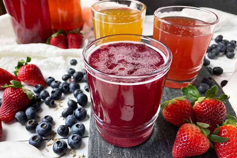 Horizontal image of three glasses filled with assorted drinks on a dark slate next to fresh fruit.