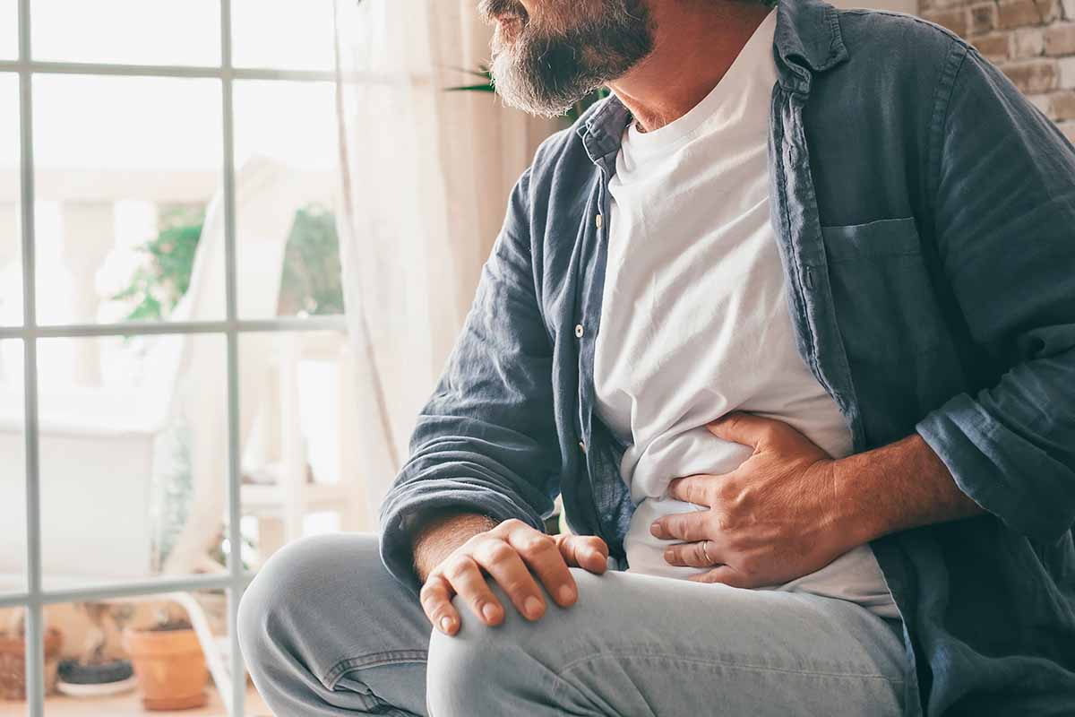 Horizontal image of a man suffering from stomach issues while he sits near a window.
