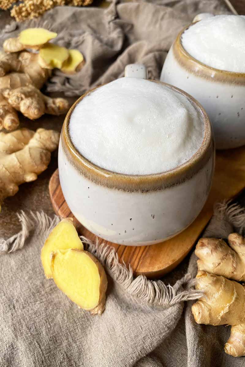 Vertical image of two mugs filled with a foamy hot drink on a wooden cutting board next to tan towels and sliced yellow roots.
