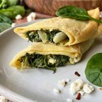 Horizontal image of a halved and stack thin pancake with a greens, onion, and cheese stuffing on a plate next to a wooden serving platter and green leaves.