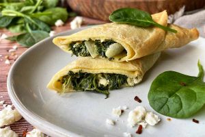 Horizontal image of a halved and stack thin pancake with a greens, onion, and cheese stuffing on a plate next to a wooden serving platter and green leaves.