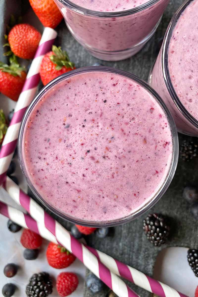 Vertical top-down image of cups filled with a bright purple shake next to fresh fruit and straws.