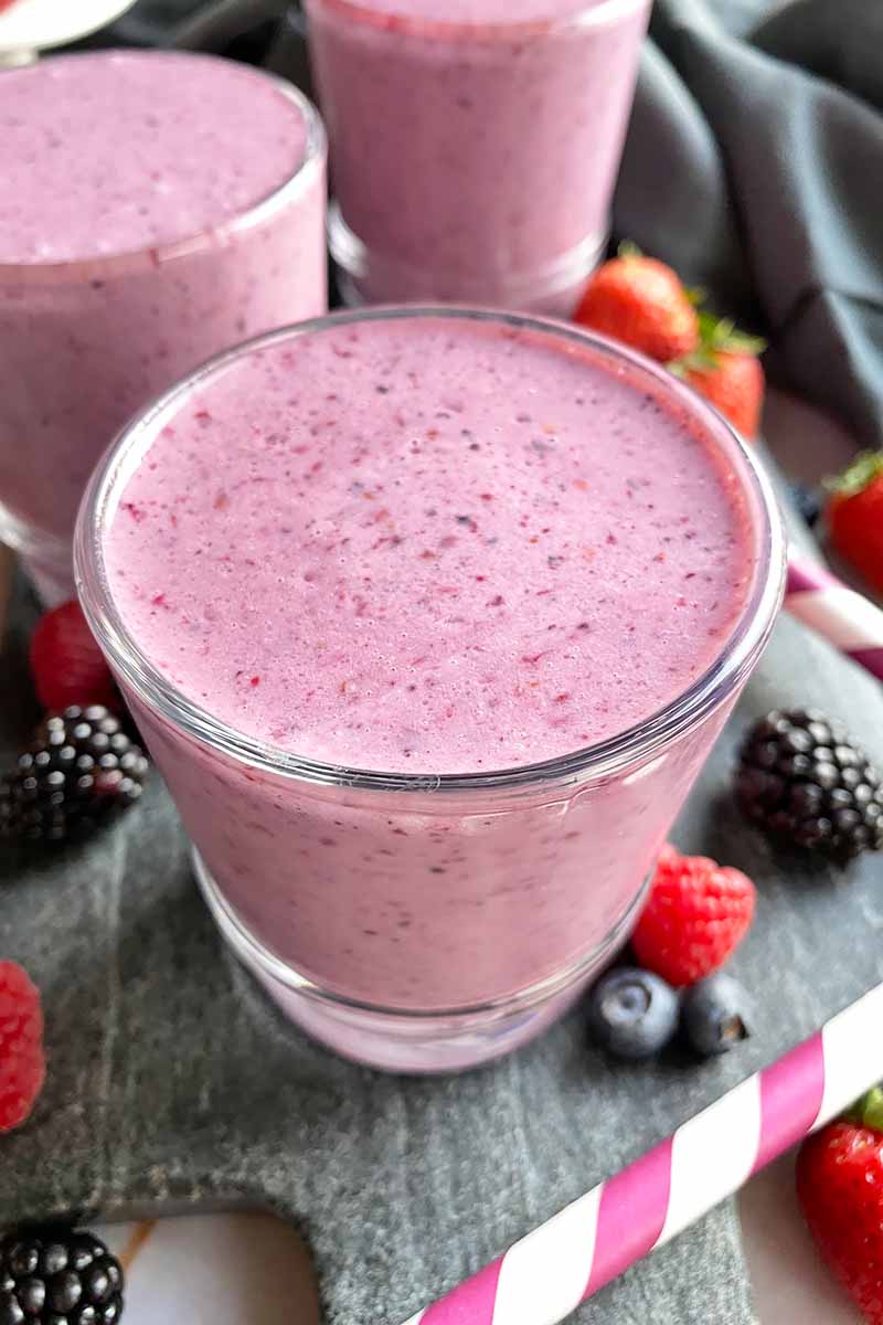 Vertical image of three glasses filled with a mixed fruit shake on a slate next to a straw.