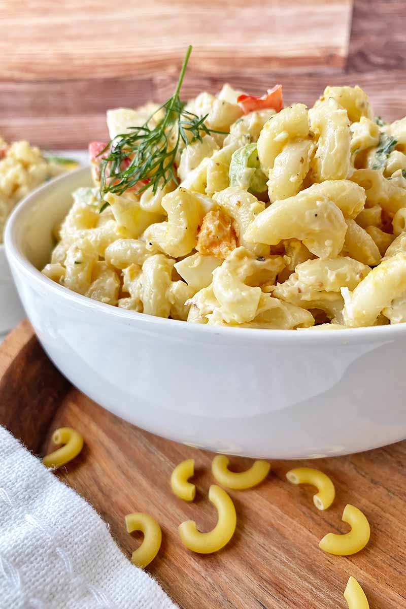 Vertical close-up image of a large bowl filled with a mixed vegetable chilled pasta dish garnished with dill on a wooden platter.