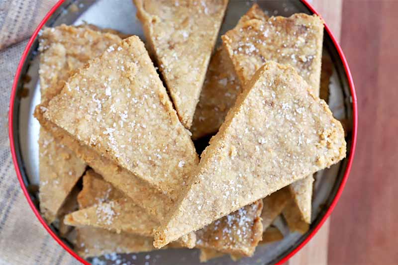 Overhead shot of a red and silver metal cookie tin filled with triangular pieces of brown butter shortbread, on a wood surfaced topped with a cloth.