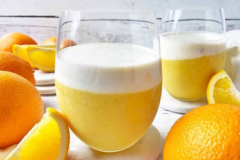Horizontal image of glasses filled with a bright shake topped with a vanilla layer next to fresh fruit.